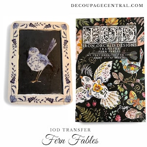May include: A framed artwork featuring a blue bird with floral patterns on a black background. The frame is cream-colored with floral and leaf designs. Next to it is a package of Iron Orchid Designs Fern Fables transfer sheets.