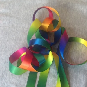 May include: A collection of rainbow-coloured satin ribbon, each ribbon is a gradient of colours, with the colours blending into each other. The ribbons are arranged in a loose knot.
