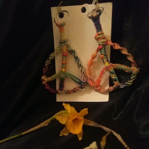 May include: Handmade macrame peace sign earrings in vibrant colors. Each earring features a peace symbol design with a loop for hanging. The earrings are accented with colorful beads. Two yellow daffodils are in the foreground.