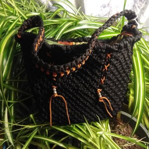May include: A black crocheted handbag with a braided handle and decorative orange stitching. The bag features orange leather accents and a textured design. The bag is set against a backdrop of green and white plants.