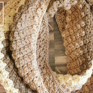 May include: A crocheted infinity scarf in brown and cream with a textured stitch pattern. The scarf is made with a thick yarn and has a warm, cozy look.