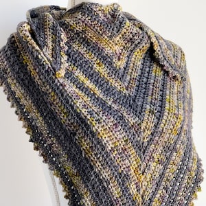 May include: A crocheted shawl in shades of gray, yellow, and brown with a scalloped edge. The shawl is draped over a white mannequin.