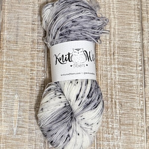 May include: A skein of speckled yarn in shades of white, gray, and black. The yarn is wrapped with a label featuring an owl logo and the text "Knit Wise fibers". The yarn is on a wooden surface.