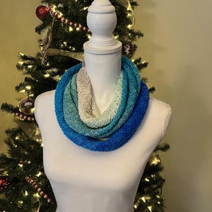 Pode incluir: Uma echarpe infinita em degradê nas cores azul, azul-turquesa e branco, feita de fio de crochê. A echarpe é exibida em um manequim branco.