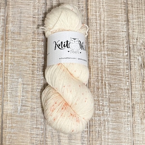 May include: A skein of off-white yarn with small orange flecks. The yarn is wrapped with a label that reads "Knit Wise Fibers" and features an owl illustration. The yarn is displayed on a light-colored wooden surface.