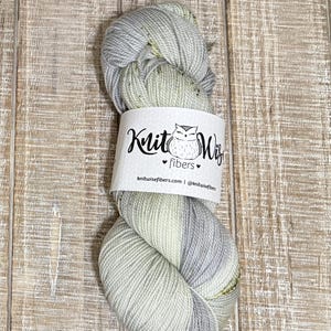 May include: A skein of Knit Wiz fibers yarn in shades of gray, green, and white. The yarn is wrapped with a white label featuring a logo of an owl and the words "Knit Wiz fibers". The background is a light brown wooden surface.