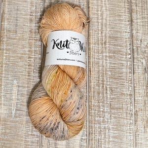 May include: A skein of Knit Wise Fibers yarn in shades of tan, peach, and speckled with dark flecks. The yarn is wrapped with a white label featuring a stylized owl and the text "Knit Wise Fibers". The background is a light brown wooden surface.