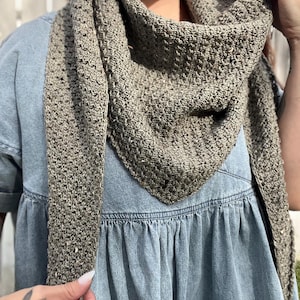 May include: A brown crocheted shawl with a textured pattern. The shawl is worn around the neck of a person wearing a denim shirt.
