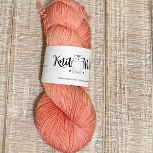 May include: A skein of coral-colored yarn with a white label featuring a stylized owl and the text "Knit Wise fibers". The yarn is tightly wound and appears soft. The background is a light brown wooden surface.