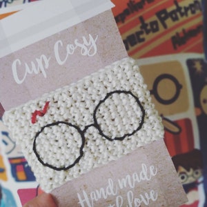 Peut inclure: Un chauffe-tasse en tricot blanc avec un motif brodé noir de lunettes et un éclair rouge. Le chauffe-tasse est présenté sur une carte brune avec le texte "Cup Cosy" et "Hand made with love".