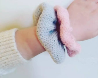May include: A knitted scrunchie in soft pastel colors, light blue and pink, is wrapped around a wrist. The scrunchie is made of a textured yarn, adding visual interest. The background is a plain white surface.