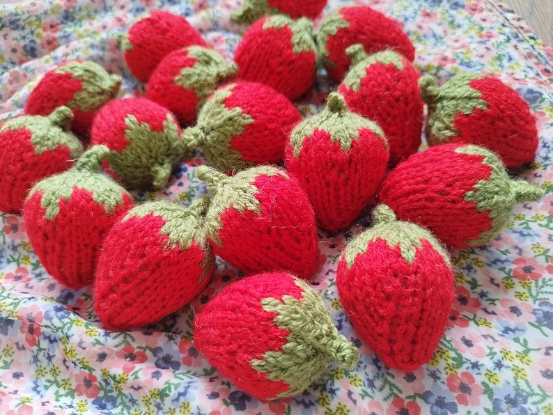 May include: A collection of knitted strawberries in various shades of red and green. The strawberries are arranged in a cluster on a floral fabric background.