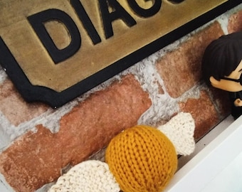 Könnte beinhalten: Ein gestrickter goldener Schnatz mit cremefarbenen Flügeln liegt auf einer weißen Oberfläche. Der Schnatz befindet sich vor einer Backsteinmauer und einem Schild mit der Aufschrift "DIAGON". Eine Figur eines Charakters mit Brille ist sichtbar.