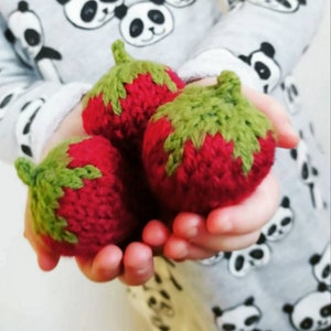 May include: Three knitted strawberries in a child's hand. The strawberries are red with green leaves and are made with a soft yarn.