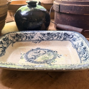 May include: A rectangular ceramic baking dish featuring a cream-colored base with a blue floral transferware pattern. The design includes a central circular motif surrounded by a decorative border of flowers and lattice details along the rim.