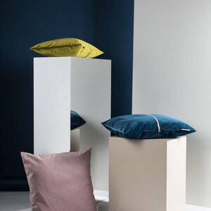 May include: Three decorative throw pillows on white cube pedestals. The pillows are a mustard yellow, a teal blue, and a dusty rose color. The pillows are arranged on a white surface against a dark blue wall.