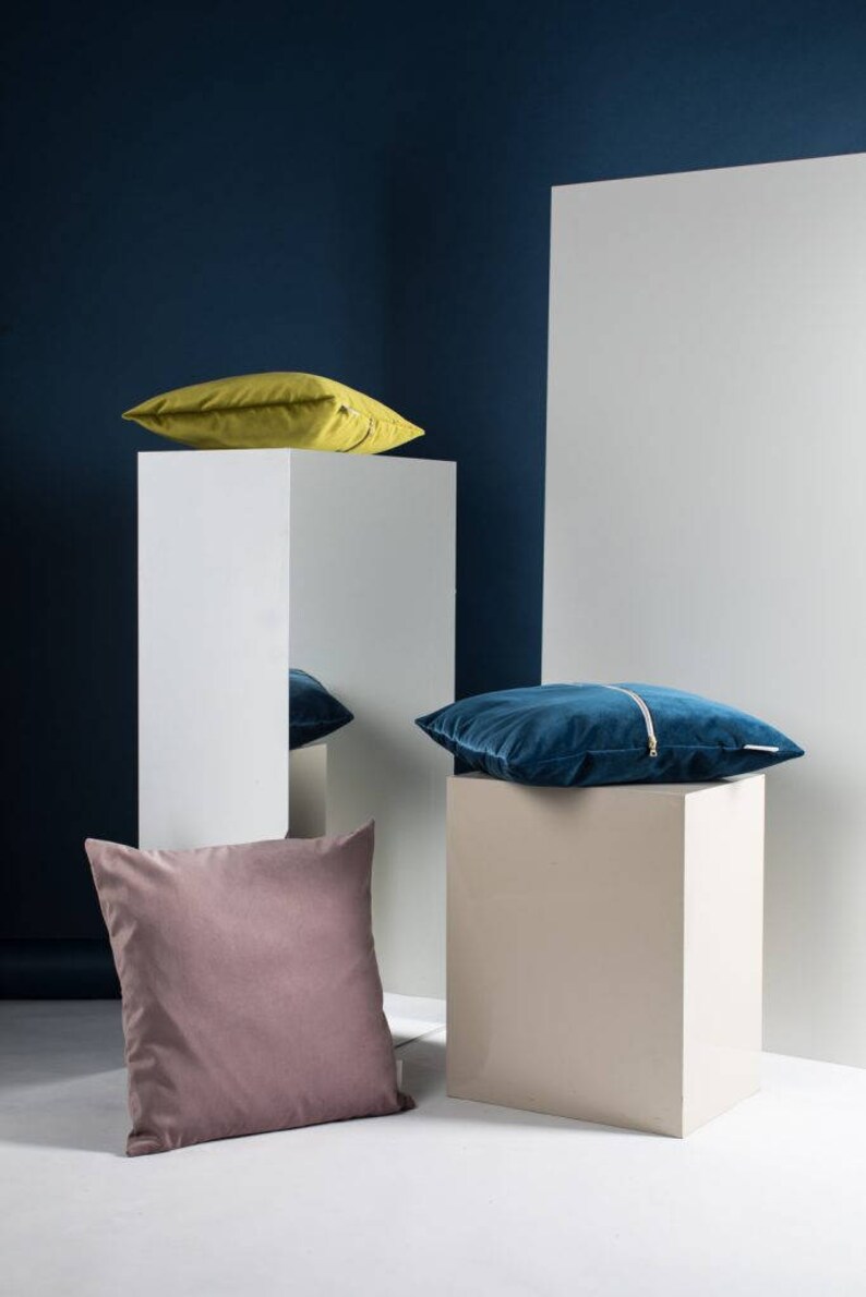 May include: Three decorative throw pillows on white cube pedestals. The pillows are a mustard yellow, a teal blue, and a dusty rose color. The pillows are arranged on a white surface against a dark blue wall.