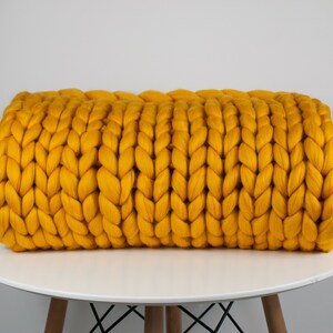 May include: A large, chunky knit throw blanket in a bright yellow color. The blanket is rolled up and sitting on a white table.