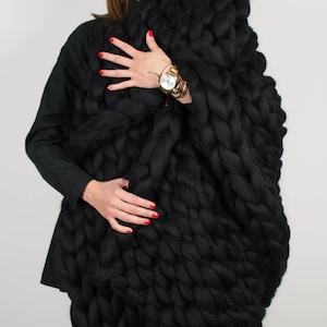 May include: A woman wearing a black oversized chunky knit blanket scarf. The scarf is made of thick yarn and has a cable knit pattern.