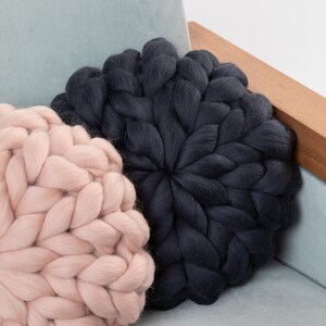 May include: Two large, round knitted throw pillows. One is a light pink color and the other is a dark charcoal gray. The pillows are sitting on a light blue couch.