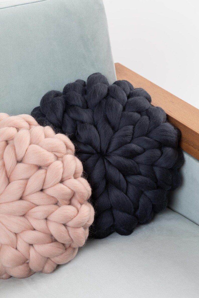 May include: Two large, round knitted throw pillows. One is a light pink color and the other is a dark charcoal gray. The pillows are sitting on a light blue couch.