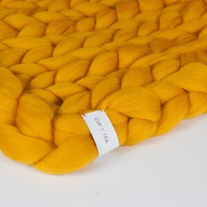 May include: A close-up of a yellow chunky knit throw blanket with a white tag that says "CUP + TEA".