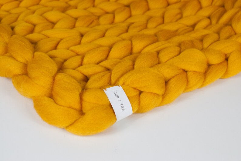 May include: A close-up of a yellow chunky knit throw blanket with a white tag that says "CUP + TEA".