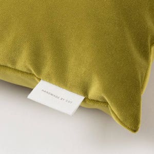 May include: A close-up of a mustard yellow velvet pillow with a white label that reads "HANDMADE BY COT".