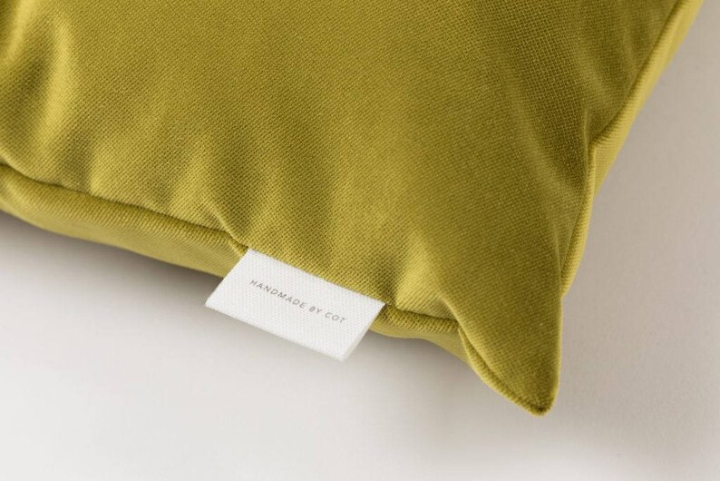 May include: A close-up of a mustard yellow velvet pillow with a white label that reads "HANDMADE BY COT".