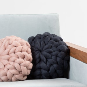 May include: Two round knitted throw pillows on a light blue couch. One pillow is a light pink color and the other is dark gray.