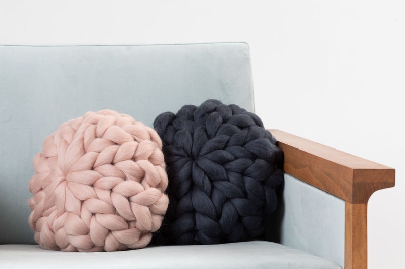 May include: Two round knitted throw pillows on a light blue couch. One pillow is a light pink color and the other is dark gray.