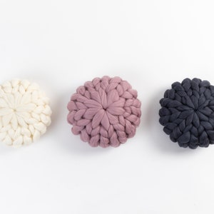 May include: Three round knitted cushions in white, pink, and dark gray. The cushions are made of thick yarn and have a textured, puffy appearance.