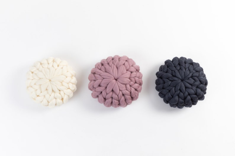 May include: Three round knitted cushions in white, pink, and dark gray. The cushions are made of thick yarn and have a textured, puffy appearance.
