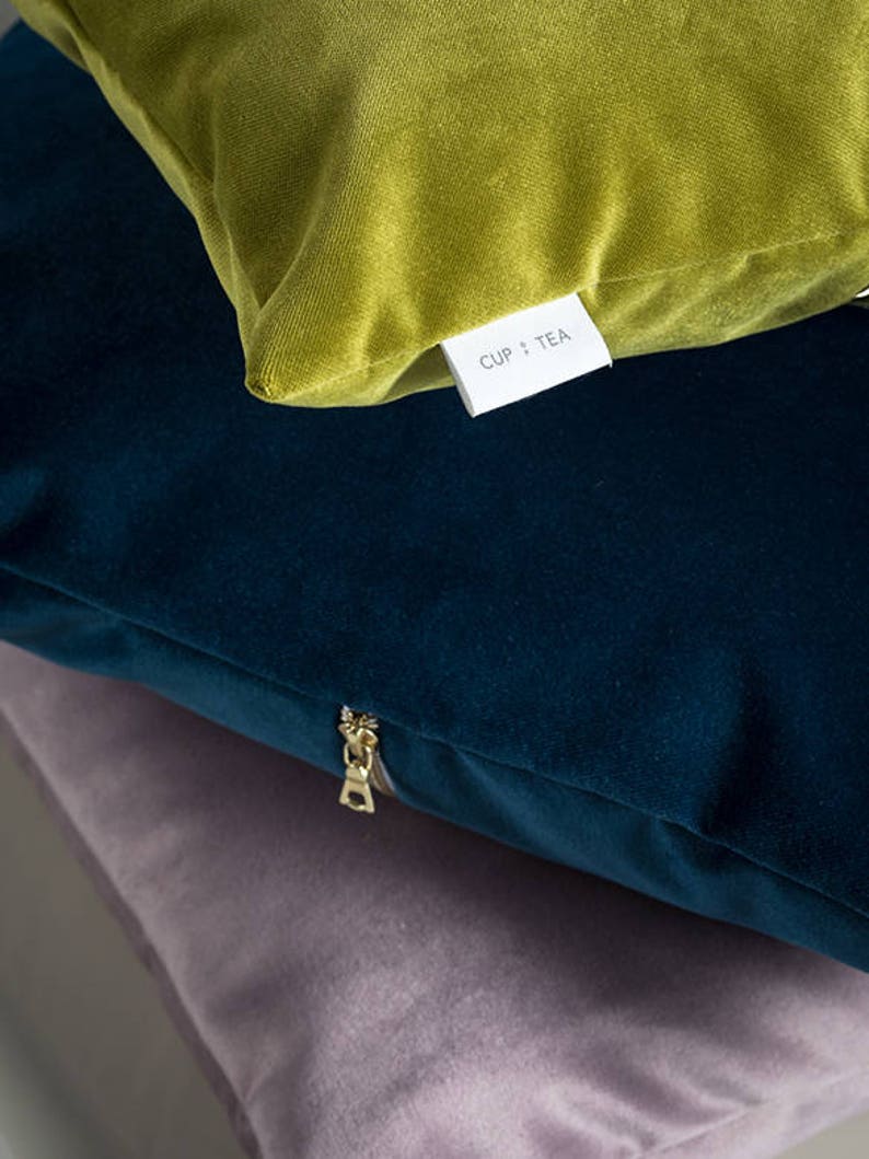 May include: Three velvet pillow covers stacked on top of each other. The top cover is a bright green, the middle cover is a dark blue, and the bottom cover is a light purple. The green cover has a white label that says "CUP + TEA". The blue cover has a gold zipper.