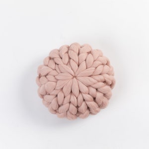 May include: A round, blush-colored knitted pillow with a unique, textured design. The pillow is made of soft, fluffy yarn and has a circular shape.