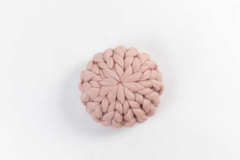 May include: A round, blush-colored knitted pillow with a unique, textured design. The pillow is made of soft, fluffy yarn and has a circular shape.