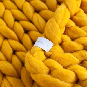 May include: A close-up of a chunky knit throw blanket in a bright yellow color. The blanket is made with thick yarn and has a soft, fuzzy texture. A white tag with the text "Handmade by Cott" is attached to the blanket.