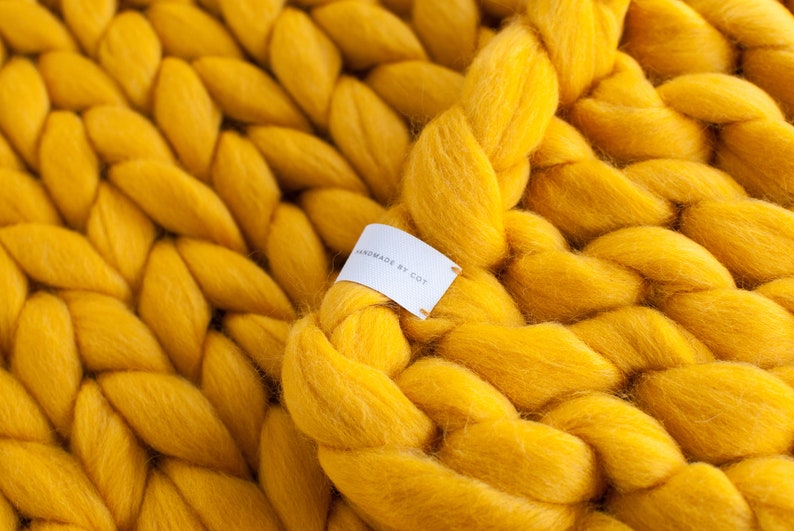 May include: A close-up of a chunky knit throw blanket in a bright yellow color. The blanket is made with thick yarn and has a soft, fuzzy texture. A white tag with the text "Handmade by Cott" is attached to the blanket.
