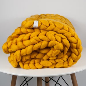 May include: A mustard yellow chunky knit throw blanket folded on a white table. The blanket is made of thick, soft yarn and has a braided texture.