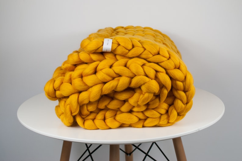 May include: A mustard yellow chunky knit throw blanket folded on a white table. The blanket is made of thick, soft yarn and has a braided texture.