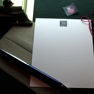 May include: A blank white card with a small, dark gray monogram in the upper right corner. A blue pen is lying on the card, and a red string is tied around the card. The card is on a brown envelope.