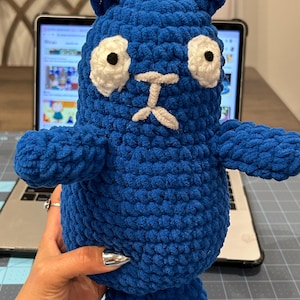 May include: A handmade blue crochet stuffed animal with white eyes and a light gray mouth. The plush toy has small arms, ears, and legs. The toy is being held in front of a laptop.