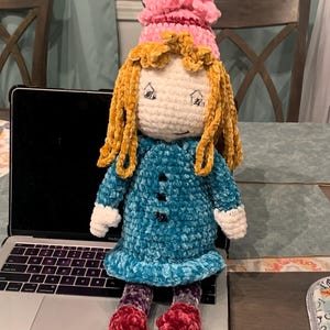 May include: A handmade crochet doll with a white face, yellow hair, and a pink hat. The doll wears a blue dress with black buttons and red shoes. The doll is sitting on a laptop.