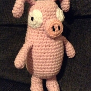 May include: A handmade pink crochet pig stuffed animal with white eye accents and black pupils. The pig has a round snout, small ears, and black hooves. This whimsical pig is a unique handmade item.
