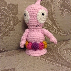 May include: A handmade pink crochet alien doll with white eyes and a small smile. The doll has a purple pom-pom on its head and a belt of colorful pom-poms. The doll is sitting on a patterned surface.