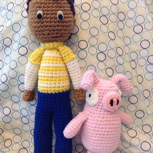 May include: A handmade crochet doll featuring a brown-skinned figure with dark hair, a blue cap, a yellow and white striped shirt, and blue pants, standing next to a small, light pink crochet pig with black snout details and white eye patches.