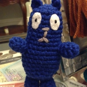 May include: A handmade, blue crochet creature with white eyes and a gray nose and mouth. The plush toy has small arms and legs. The background includes currency and a decorative mirror.