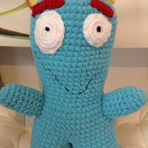 May include: A handmade, light blue crochet monster doll with white eyes, red eyebrows, yellow horns, and a stitched black smile. The plush toy has arms and legs, perfect for a child's play or room decor.