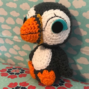 May include: A handmade crochet puffin bird with a white belly, gray back, and orange beak and feet. The puffin has large blue eyes and is set against a background of a floral pattern and a sky-blue fabric with white clouds.