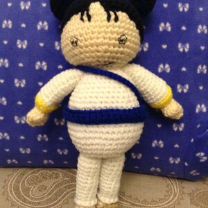May include: A handmade crochet doll with a white body, blue accents, and black hair styled in two buns. The doll has a tan face and feet, and is wearing a blue strap across the chest. The background is a blue pillow with white butterfly patterns.